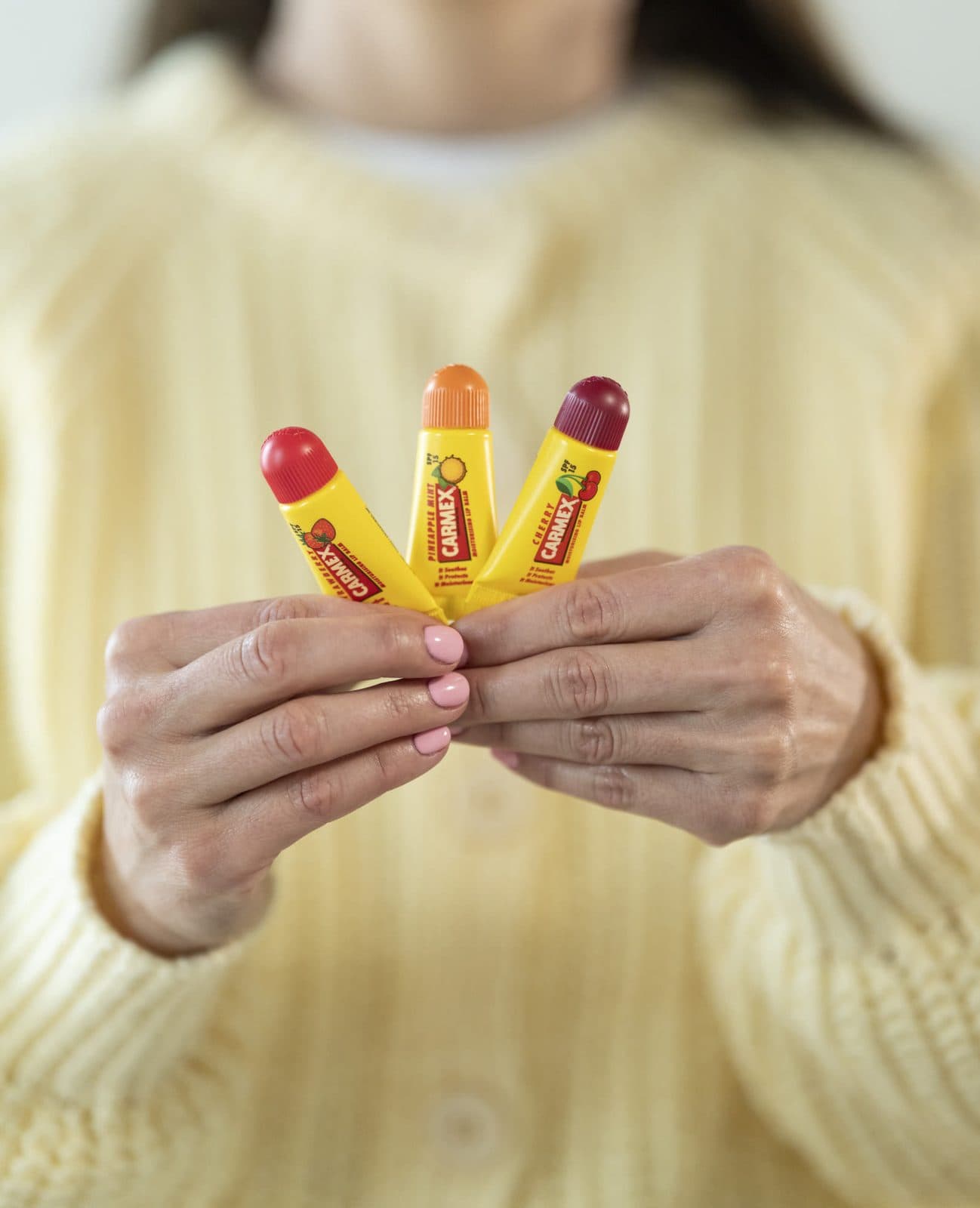 Una persona con un maglione giallo tiene tre tubetti Carmex aromatizzati