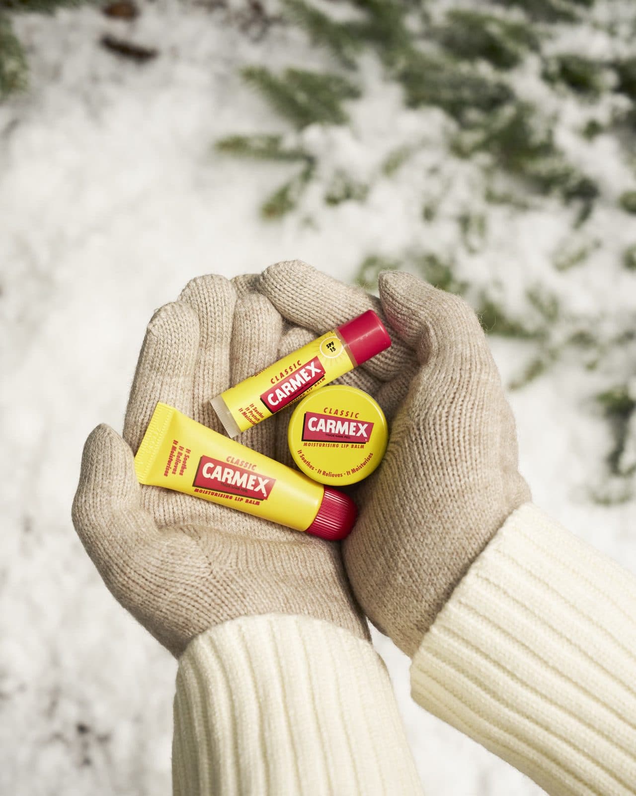 Mani con guanti di lana tengono Carmex Classic in vasetto, tubetto e stick sopra la neve